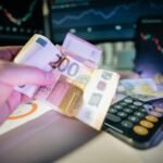 A hand holds euro notes with financial charts and a calculator, symbolizing investment and trading.