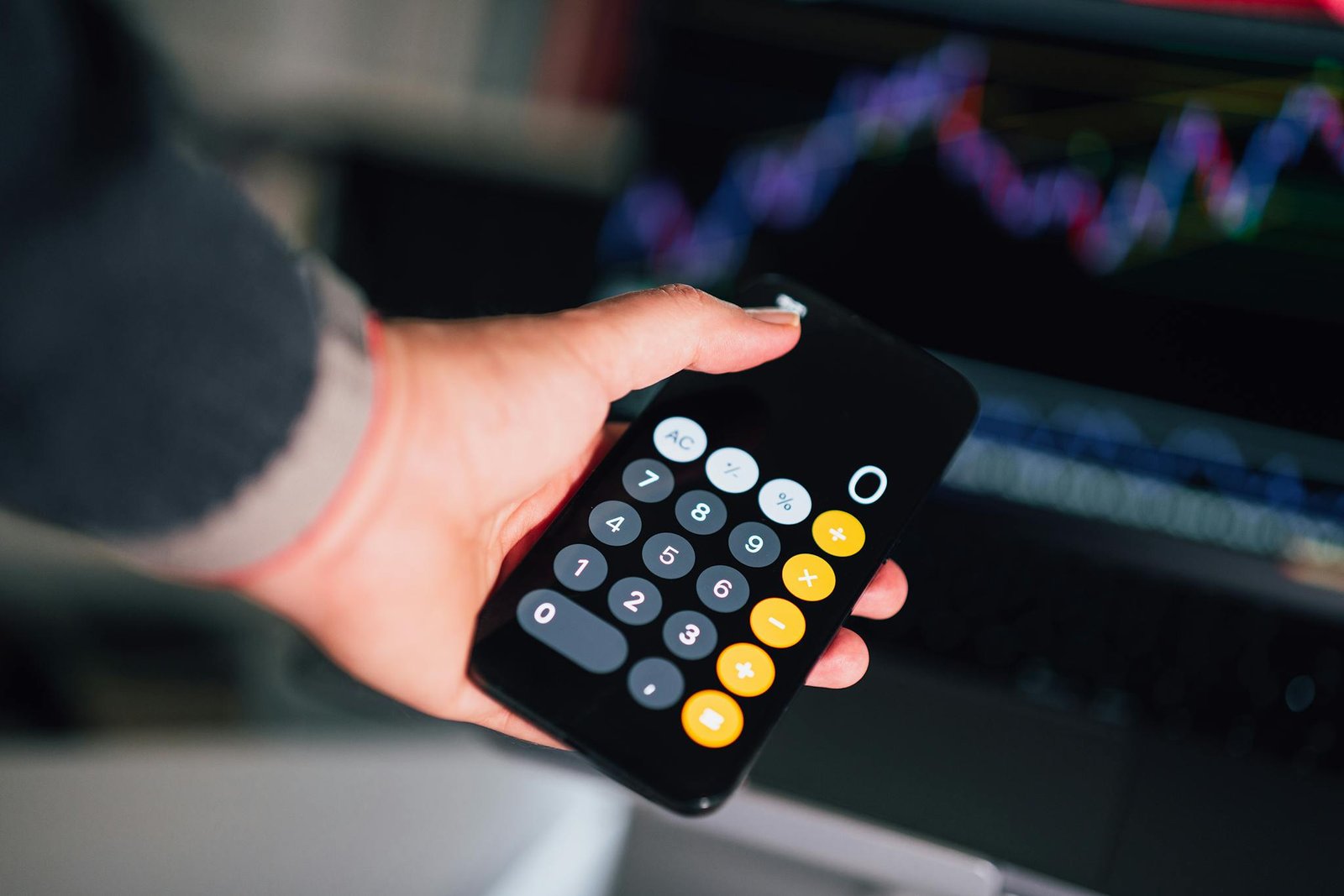 Close-up of a hand holding a smartphone calculator with financial charts on a screen in the background.